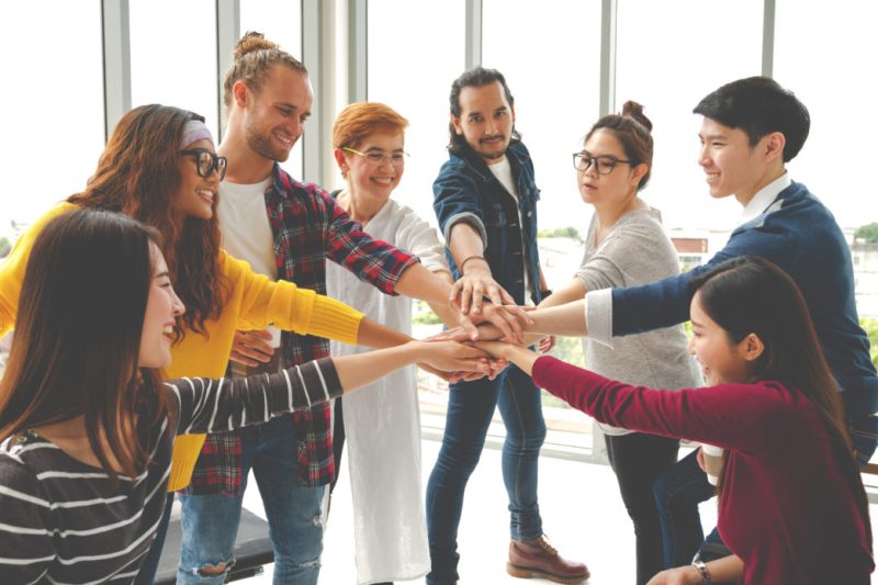 Multiethnic young team stack hands together as unity and teamwork in modern office. Diverse group togetherness collaboration or friends huddle concept. Startup business team building with soft tone.